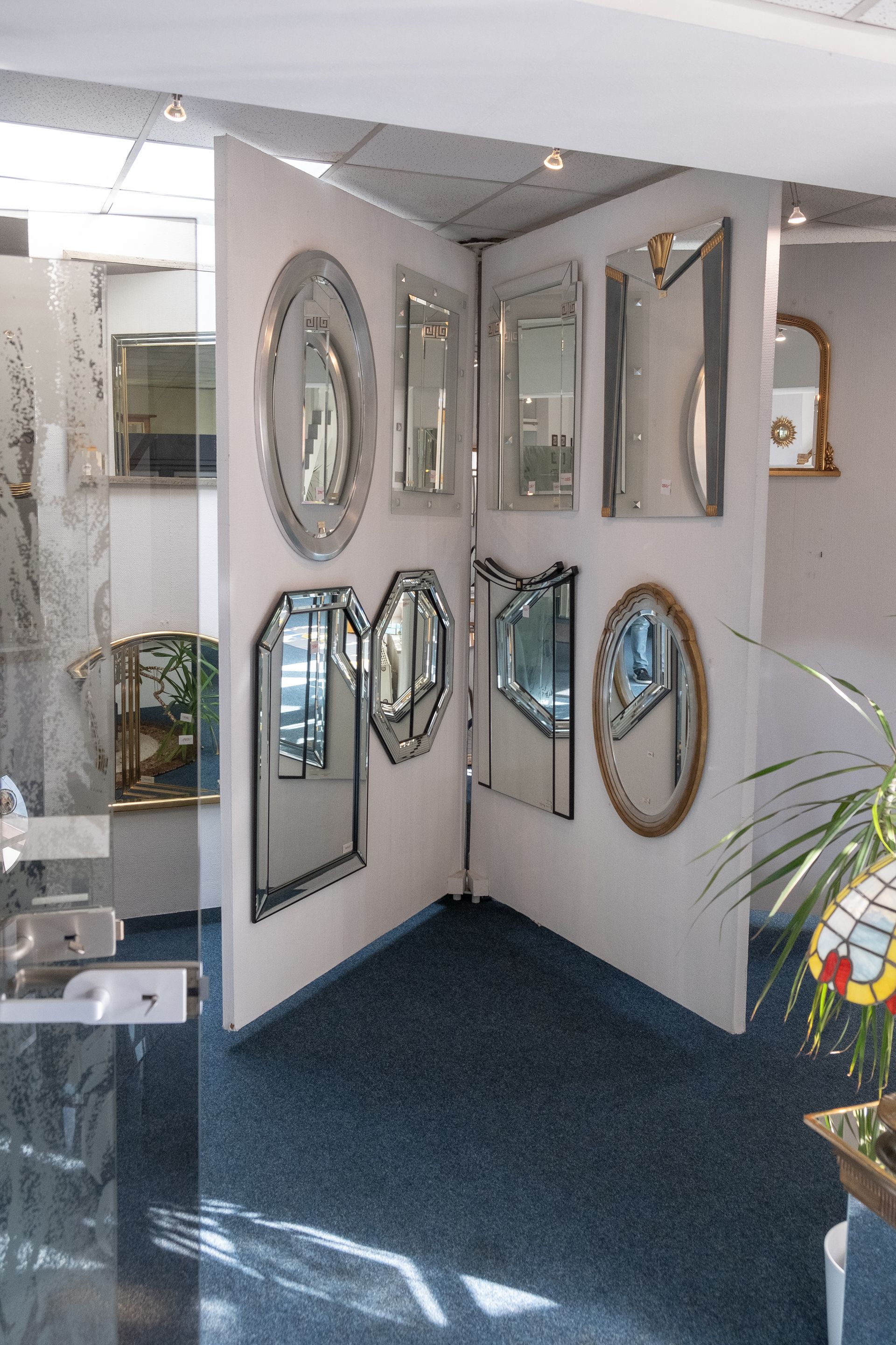 Ausgestellte Spiegel in verschiedenen Formen und Größen, angeordnet auf weißen Tafeln in einem Ausstellungsraum. Blauer Teppich, helle Beleuchtung.