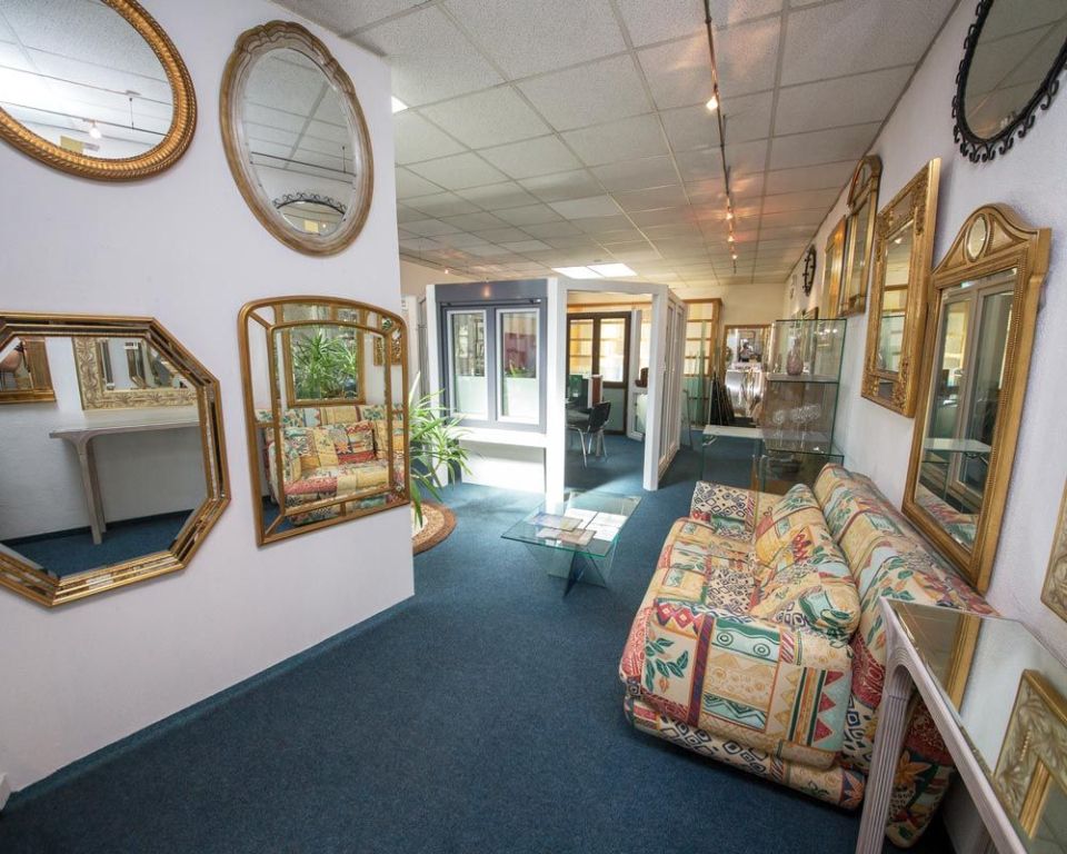 In einem Ausstellungsraum hängen Spiegel an den weißen Wänden. Auf einem blauen Teppich steht ein gemustertes Sofa.