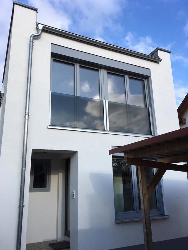 Weißes Gebäude mit grauen Fensterrahmen und einem Glasbalkon; sonniger Tag.