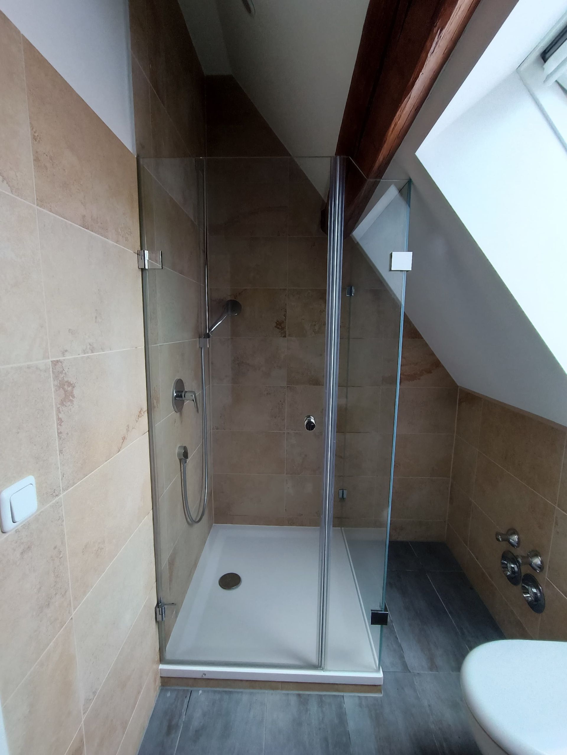 Duschkabine mit Glastüren in einem Badezimmer, schräge Decke. Beige Fliesen und eine weiße Duschwanne.