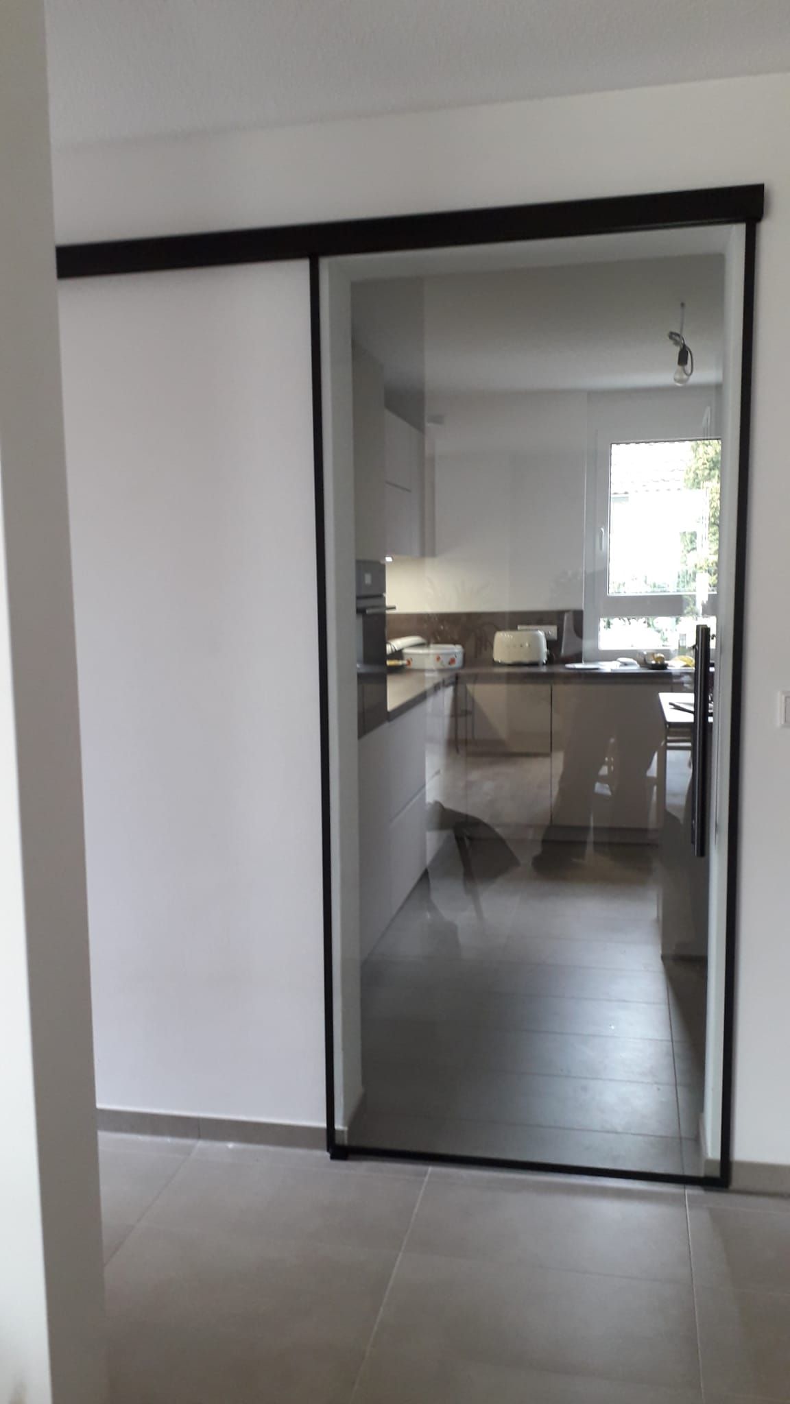 Glasschiebetür mit schwarzem Rahmen, die in eine Küche führt; die Tür ist teilweise geöffnet und gibt den Blick auf eine weiße Küche frei.