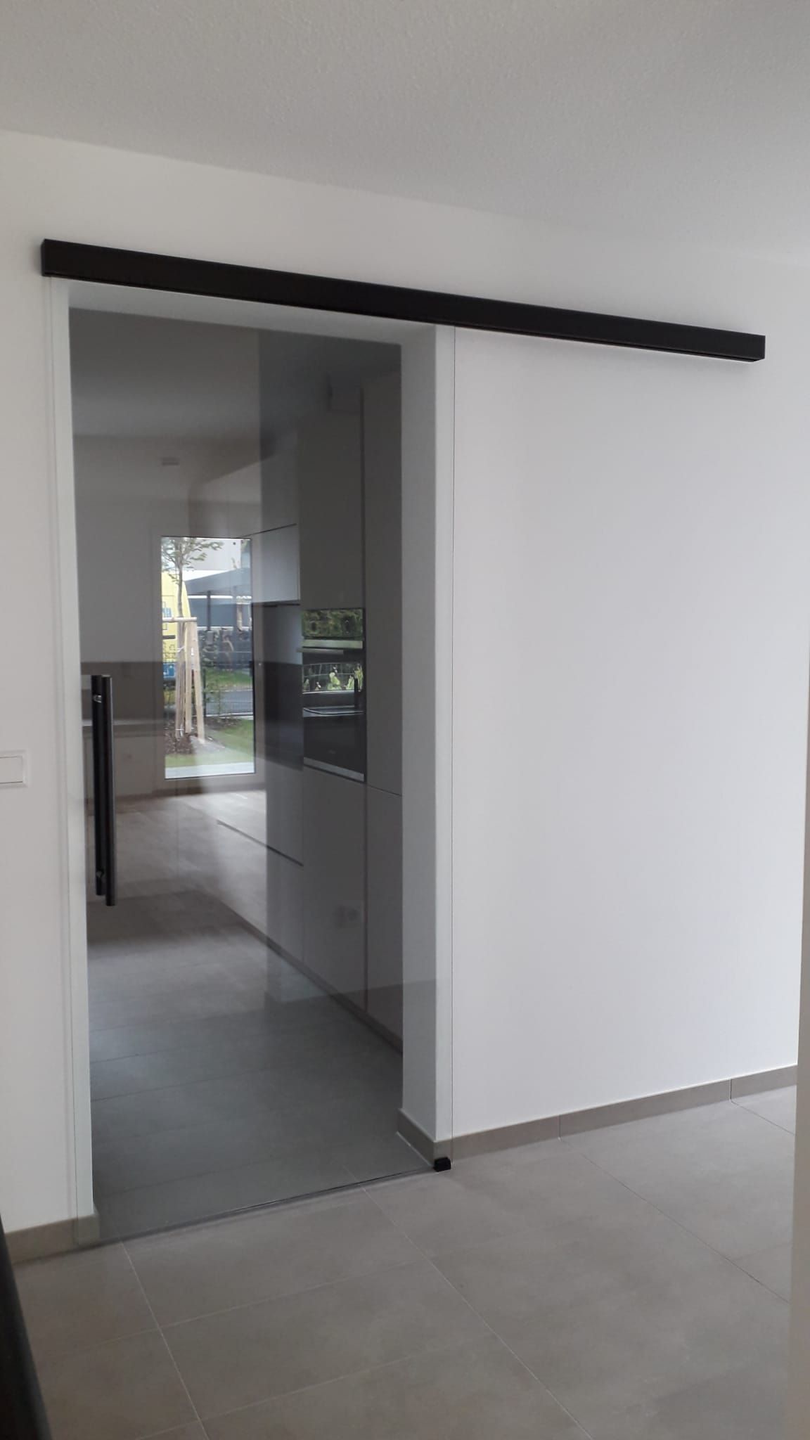 Glasschiebetür, schwarze Schiene, zwischen weißen Wänden, die den Blick auf die Küche freigibt.