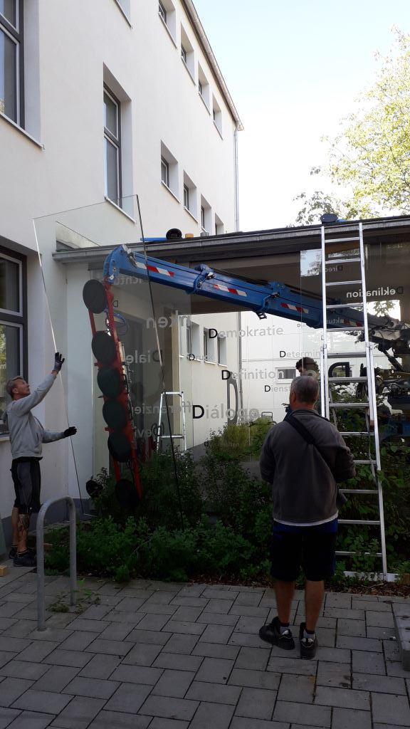 Männer arbeiten daran, ein Glasdach über einem Gebäudeeingang zu installieren. Einer auf einer Leiter, einer auf dem Boden.