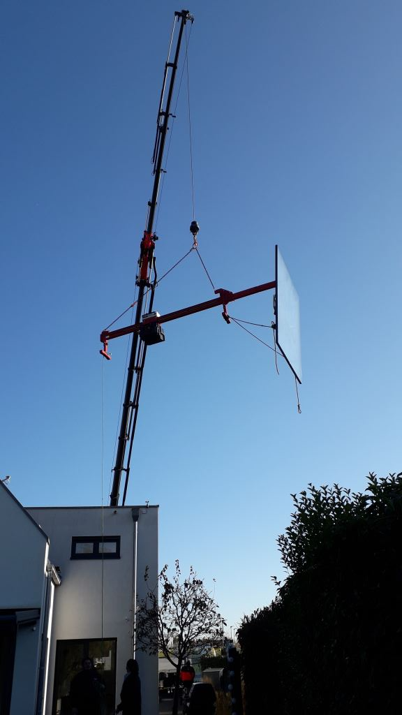 Ein Kran hebt eine große Glasscheibe, möglicherweise für ein Gebäude, vor einem blauen Himmel an.