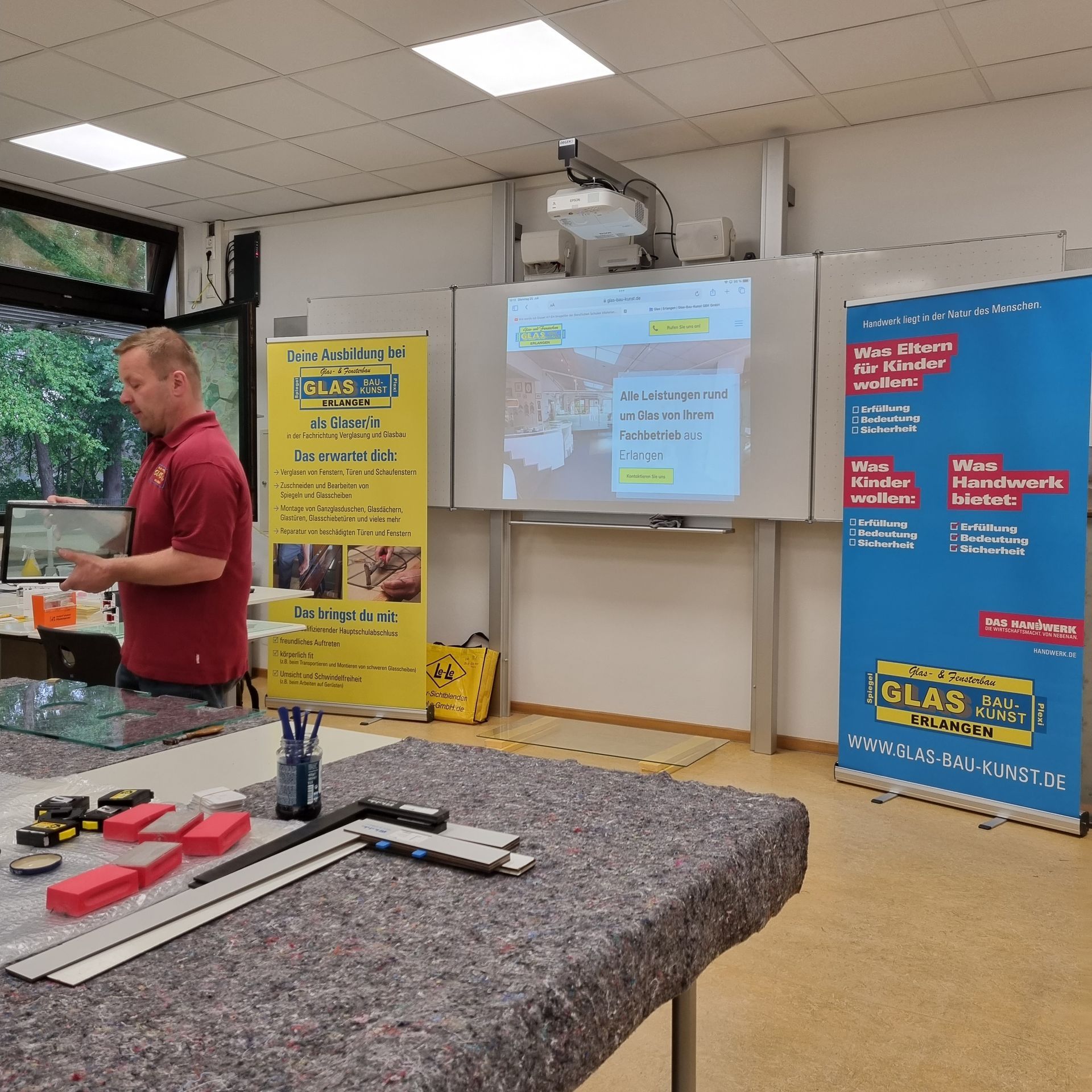 Mann hält in einem Klassenzimmer einen Vortrag über Glasarbeiten. Werkzeuge auf einem Tisch; Poster auf beiden Seiten eines Bildschirms, auf dem eine Webseite angezeigt wird.