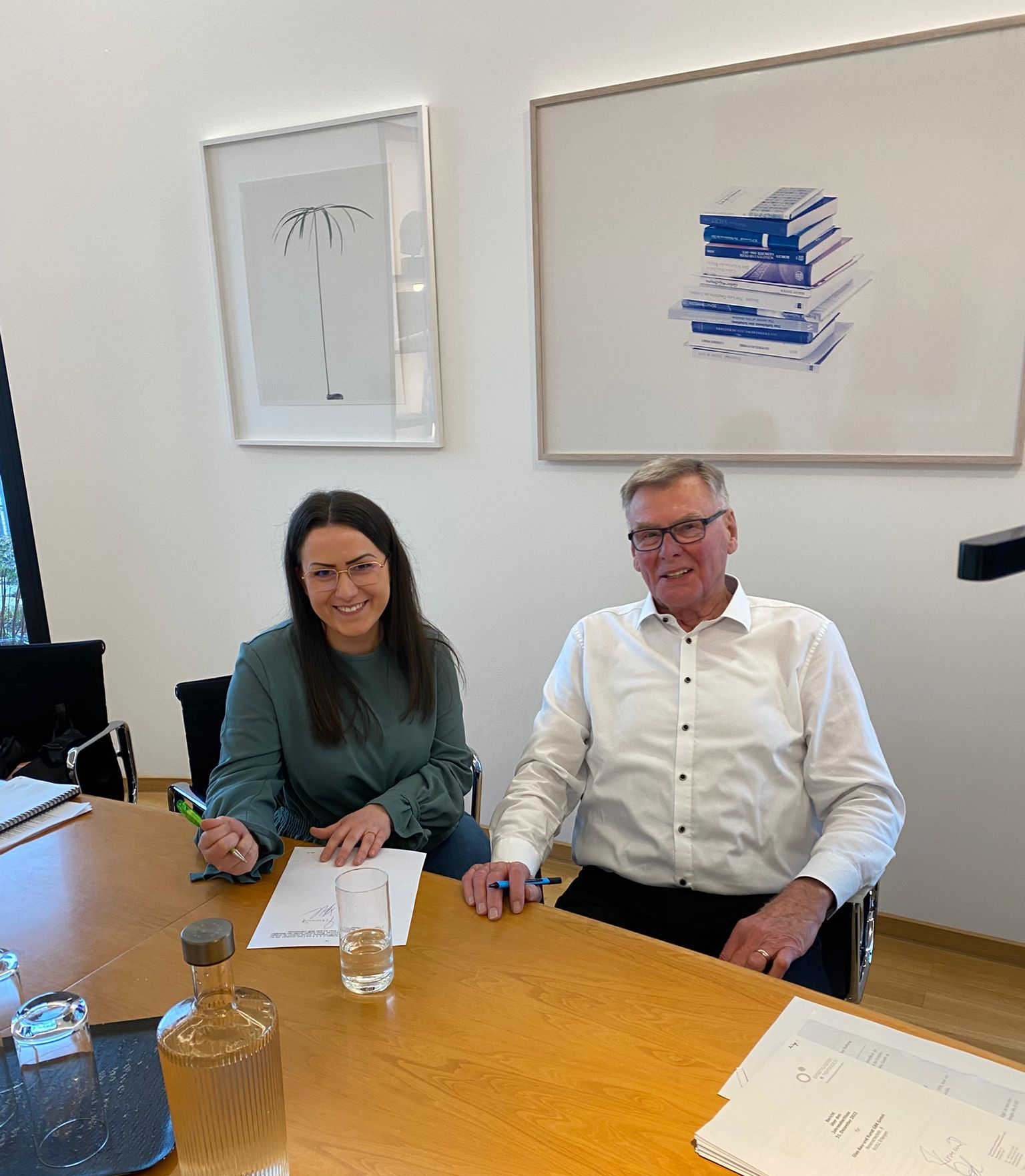 Zwei Personen, eine Frau und ein Mann, sitzen an einem Tisch. Sie scheinen Dokumente zu unterschreiben. An der Wand hängt ein Kunstwerk.