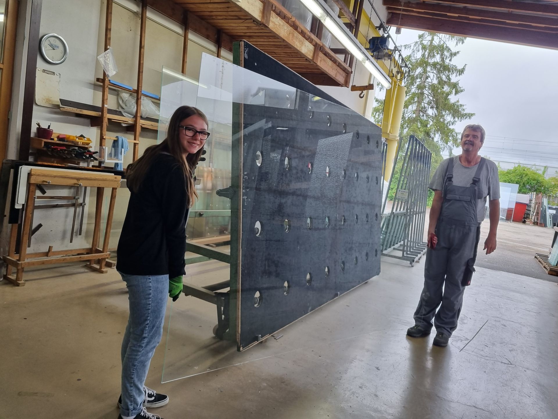 Zwei Personen handhaben in einer Werkstatt eine große Glasscheibe. Lächelnde Frau, Mann im Overall.
