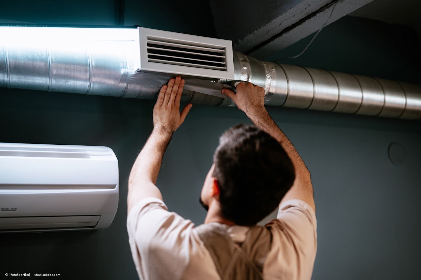 Ein Techniker inspiziert eine Deckenlüftungsöffnung neben einem Lüftungskanal und einer an der Wand montierten Klimaanlage.