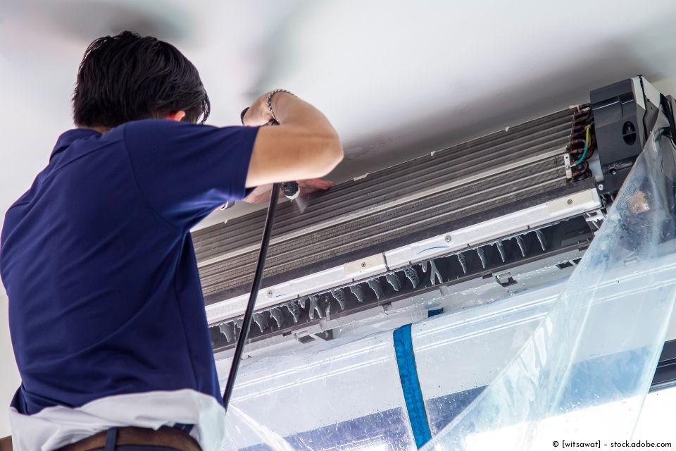 Ein Techniker reinigt in der Nähe eine Klimaanlage im Innenbereich mit einem Sprühschlauch und einer Schutzfolie.