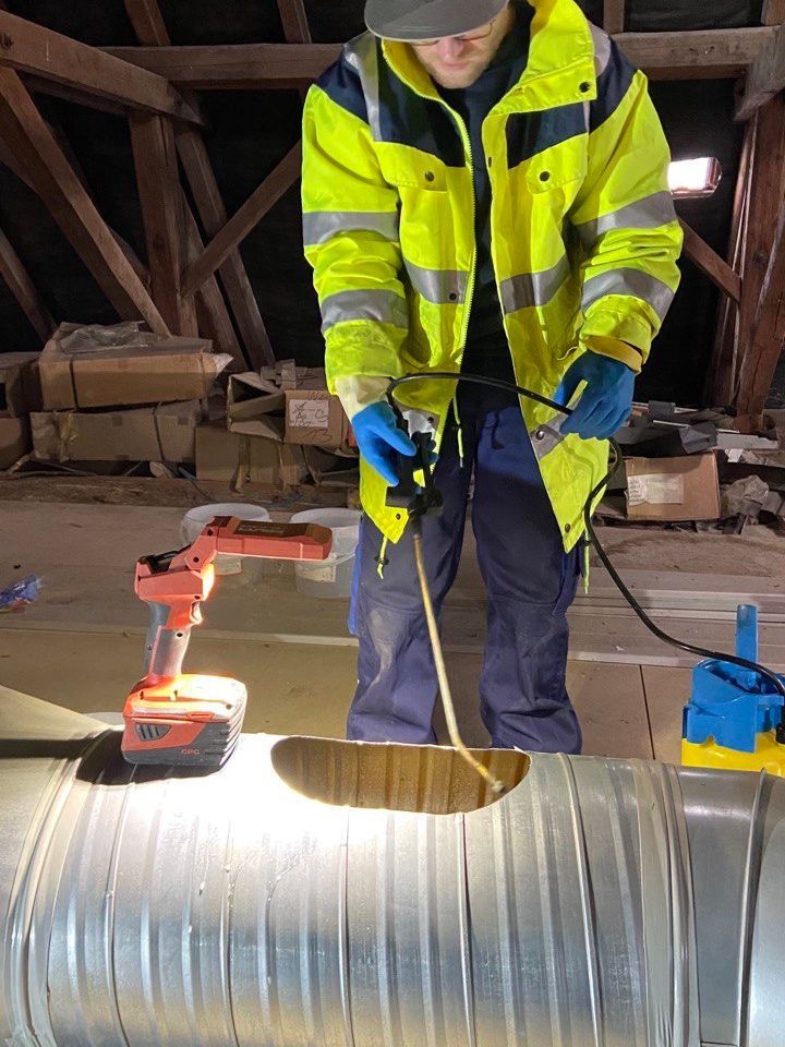Ein Arbeiter benutzt ein Elektrowerkzeug an einem Metallrohr in einem düsteren, dachbodenartigen Arbeitsbereich.