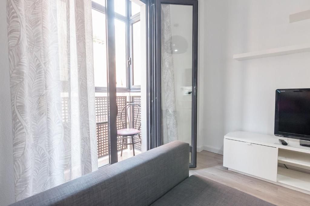 Sala de estar con sofá gris, cortinas blancas y puerta al balcón. TV sobre mueble blanco.