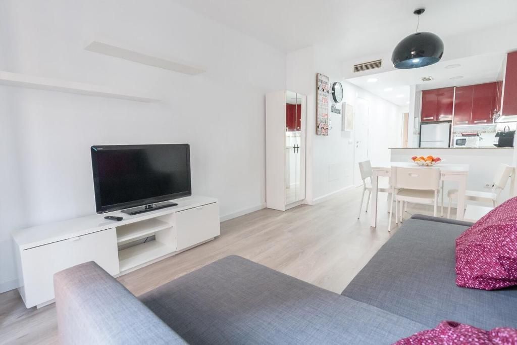 Sala de estar con sofá gris, mueble de TV blanco y cocina abierta con muebles rojos.