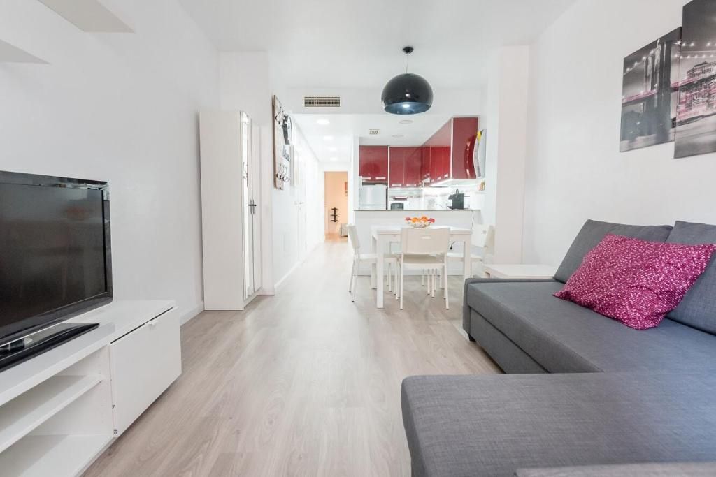 Luminosa sala de estar con sofá seccional gris, muebles blancos y una cocina visible al fondo.