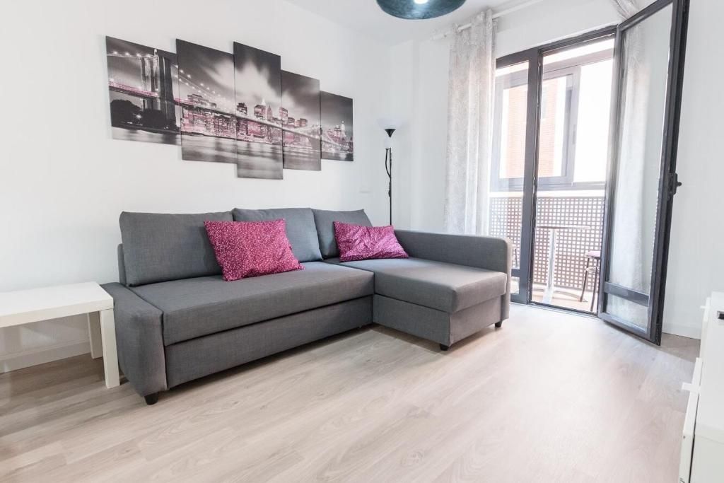 Sofá gris con cojines rosas en una sala de estar, con una obra de arte encima. Puertas del balcón abiertas.