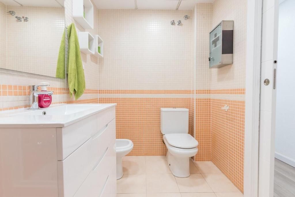 Baño con tocador blanco, inodoro y azulejos color melocotón y crema. Toalla verde colgada.