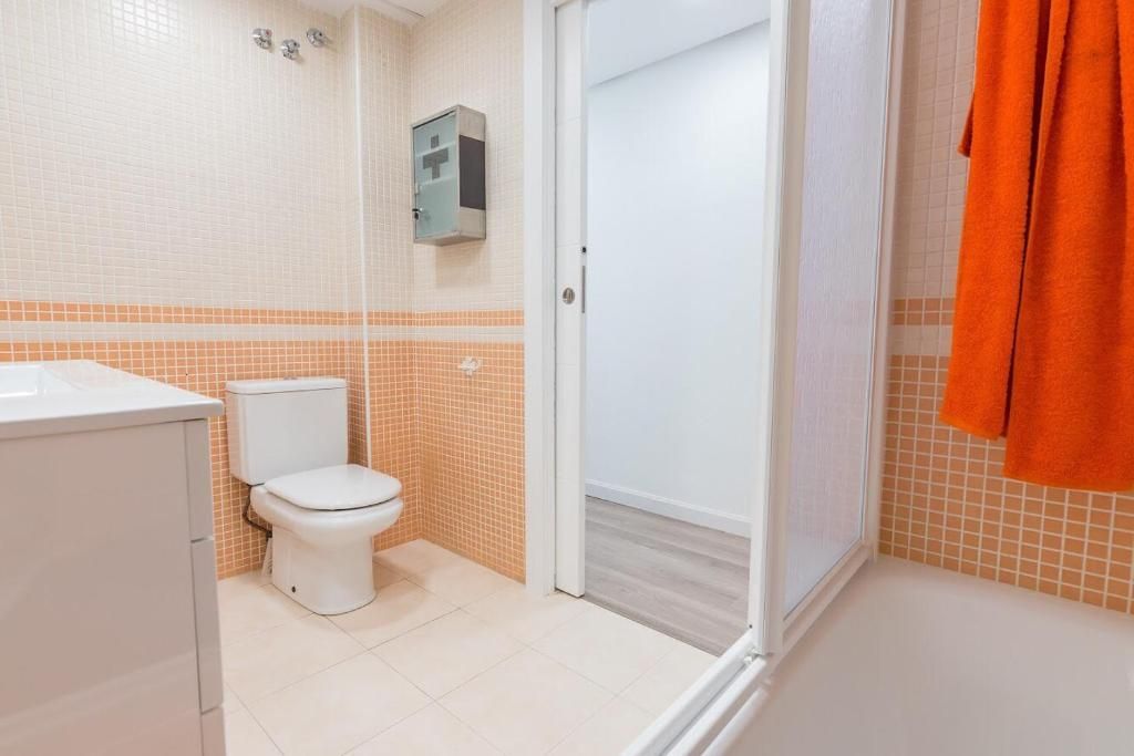 Baño con inodoro, lavabo, ducha y toalla naranja. Paredes alicatadas en blanco y naranja.