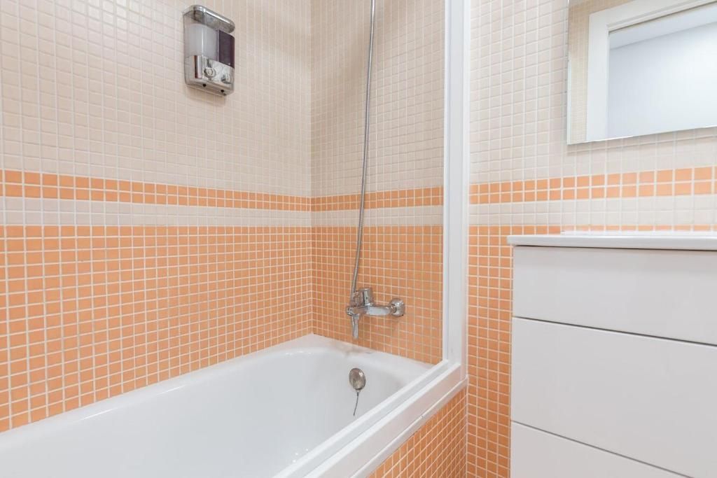 Baño con bañera blanca, paredes de azulejos color melocotón, puerta de ducha de vidrio y tocador blanco con espejo.