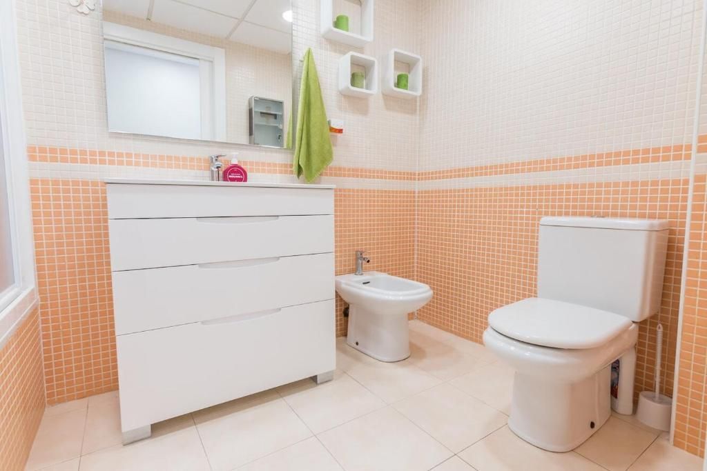 Baño con tocador blanco, inodoro y bidé. Paredes de azulejos naranjas y blancos, y una toalla verde colgada.