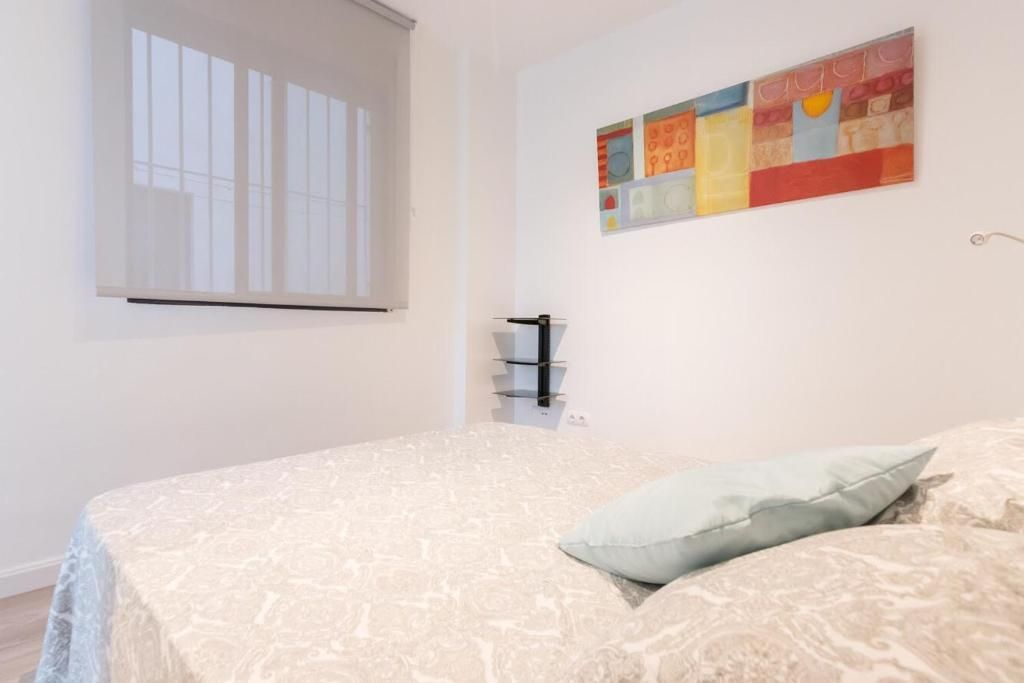 Dormitorio con cama, cuadro, pequeña estantería y ventana con persiana.