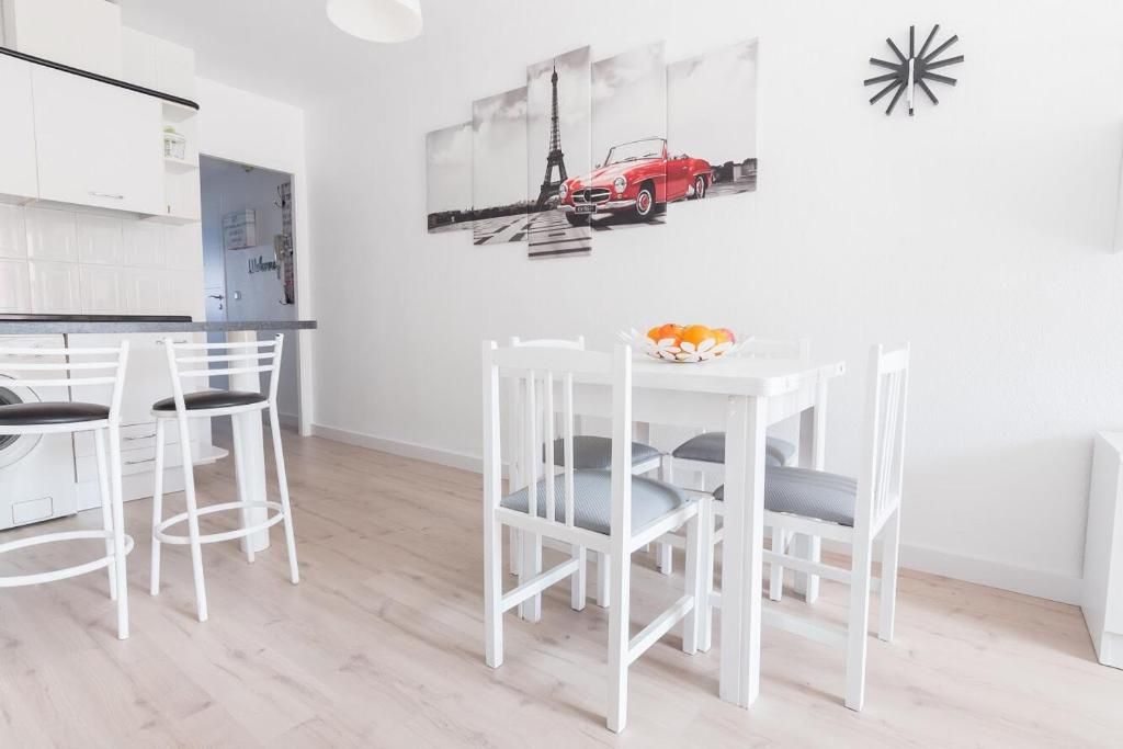 Comedor luminoso con mesa y sillas blancas, obra de arte de un coche rojo en París y un reloj negro.