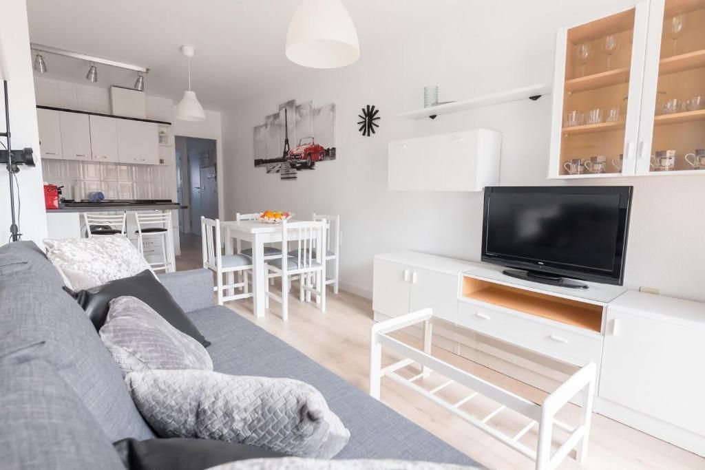 Sala de estar con muebles blancos, sofá gris, TV y comedor.