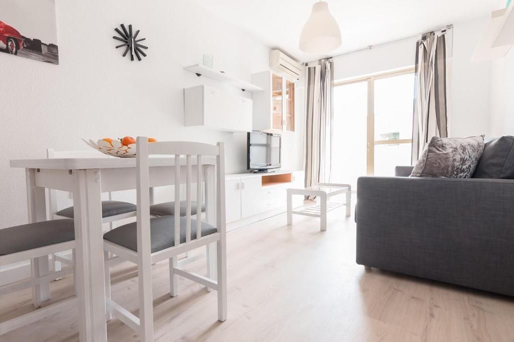 Sala de estar blanca luminosa con mesa de comedor, sofá gris y luz natural desde un balcón.