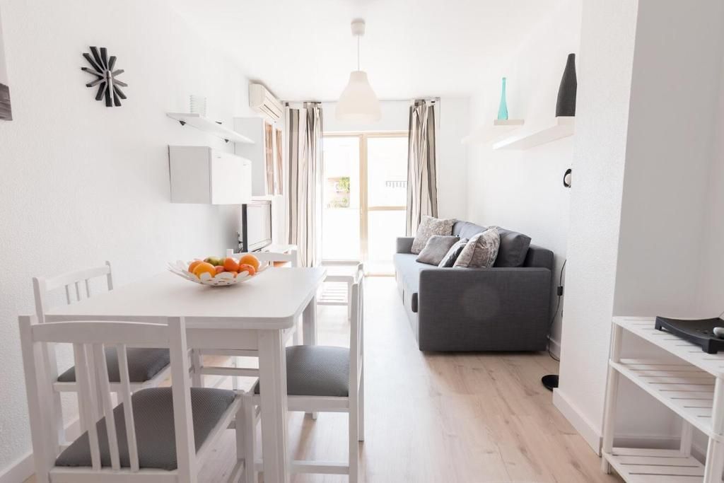 Espacio luminoso y minimalista con mesa de comedor, sofá y acceso a un balcón. Paredes blancas, suelo de madera y muebles.