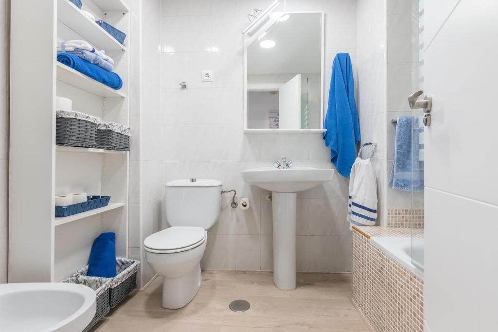 Baño blanco con inodoro, lavabo con pedestal, estanterías y bañera; toallas y detalles en azul.