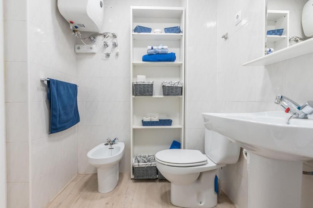 Baño blanco con inodoro, bidé, lavabo y estantería con toallas azules y cestas de almacenaje.