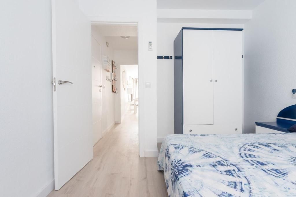 Interior de dormitorio con paredes blancas, armario, puerta abierta al pasillo, cama con funda estampada azul.