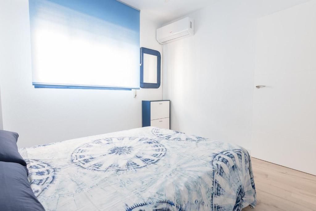 Dormitorio con cama cubierta con edredón estampado azul y blanco, ventana con pantalla azul, espejo, paredes blancas.