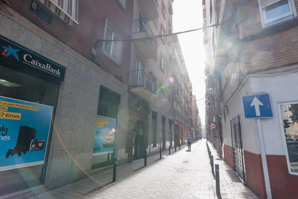 Calle estrecha de Madrid con luz solar, edificios a ambos lados, un banco y una persona caminando.