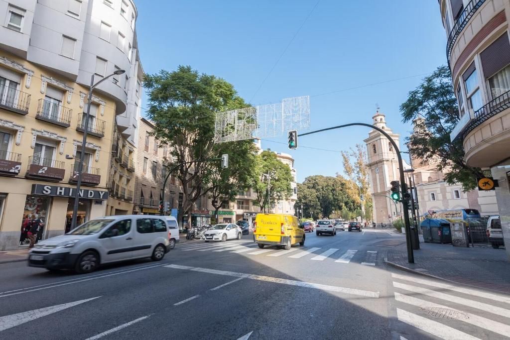 Escena callejera con vehículos, edificios y una torre de iglesia bajo un cielo azul.