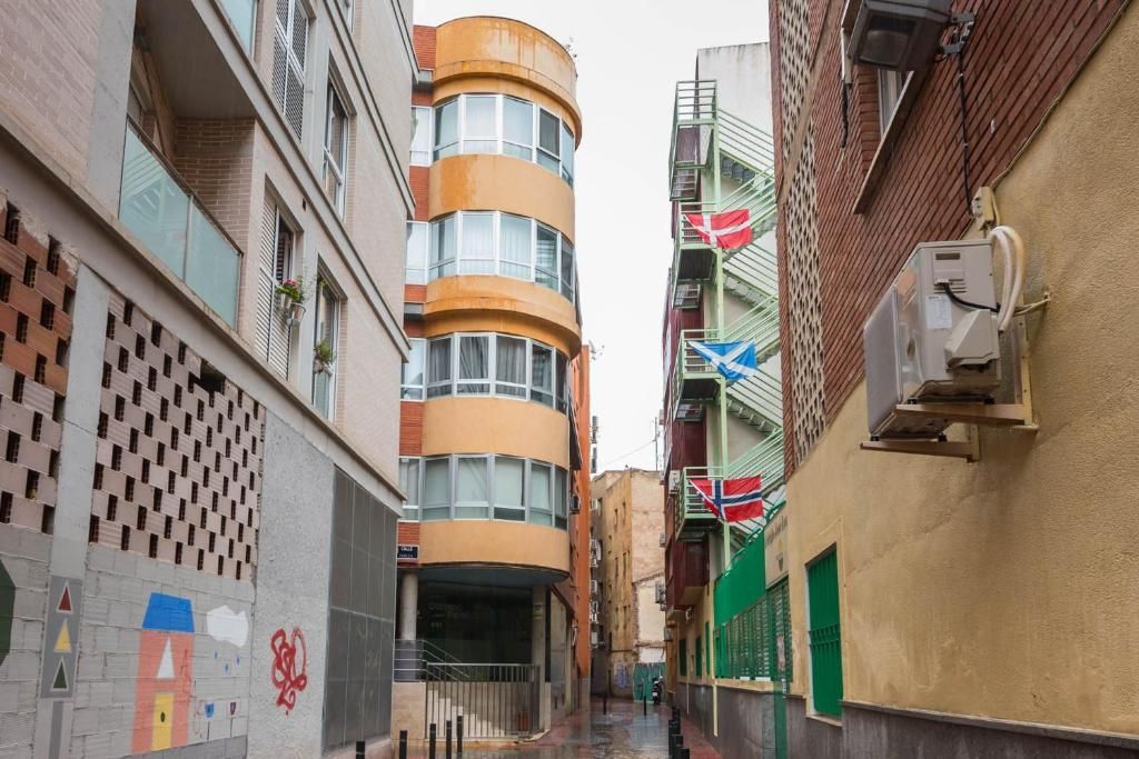 Un callejón estrecho con edificios de diferentes alturas y fachadas. Banderas cuelgan de una escalera de incendios metálica.