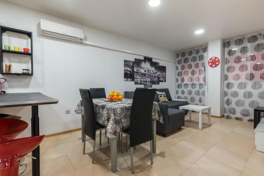 Sala de estar con mesa de comedor, sofá y taburete rojo. Paredes blancas, muebles grises y cortinas estampadas.