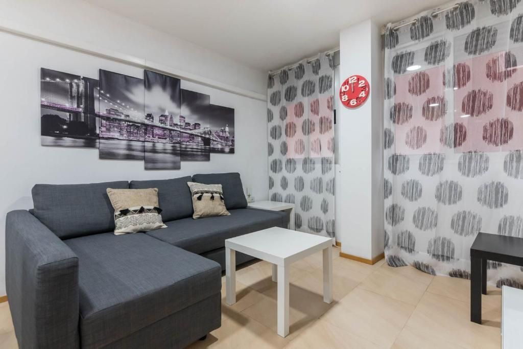 Sala de estar con sofá seccional gris, mesa blanca, arte de paisaje urbano y cortinas estampadas.