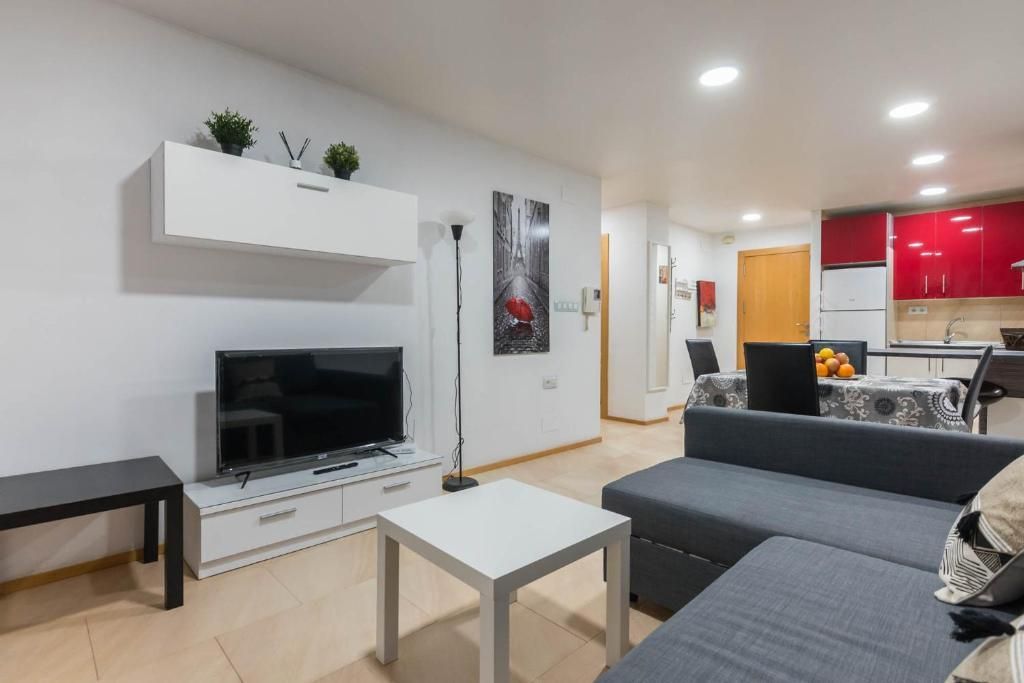 Sala de estar moderna con TV, sofá y cocina abierta. Paredes blancas, armarios rojos y muebles en tonos neutros.