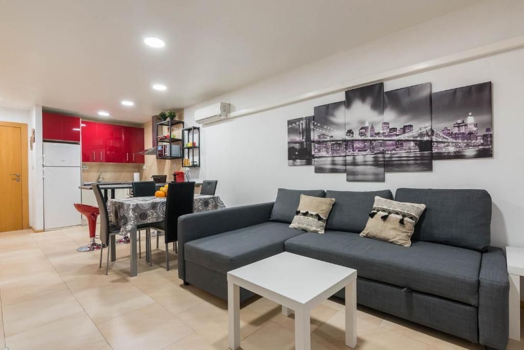 Sala de estar con sofá gris, pequeña mesa blanca y cocina abierta con muebles rojos.
