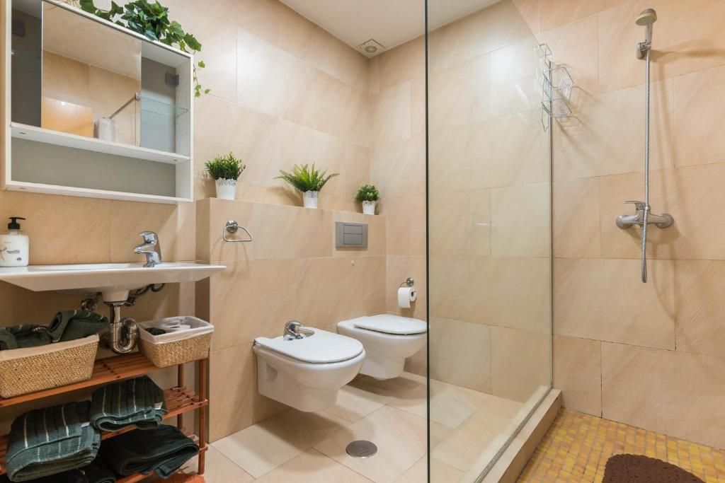 Baño con paredes de azulejos beige, ducha de cristal, inodoro, bidé y lavabo con plantas.