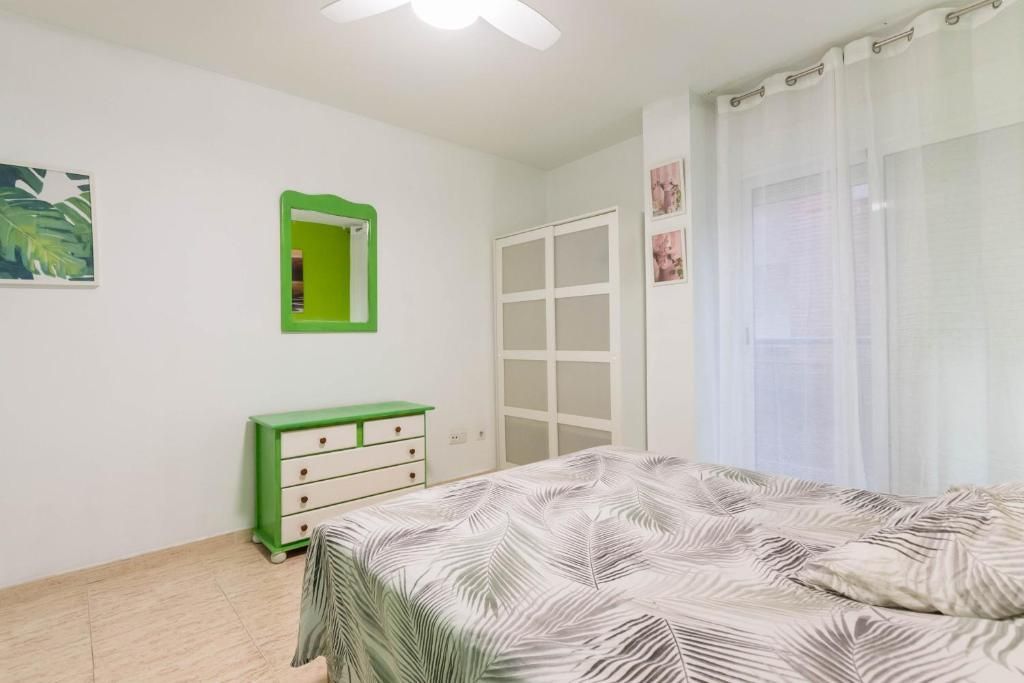 Dormitorio con paredes blancas, detalles en verde y una cama con edredón con estampado de hojas tropicales.