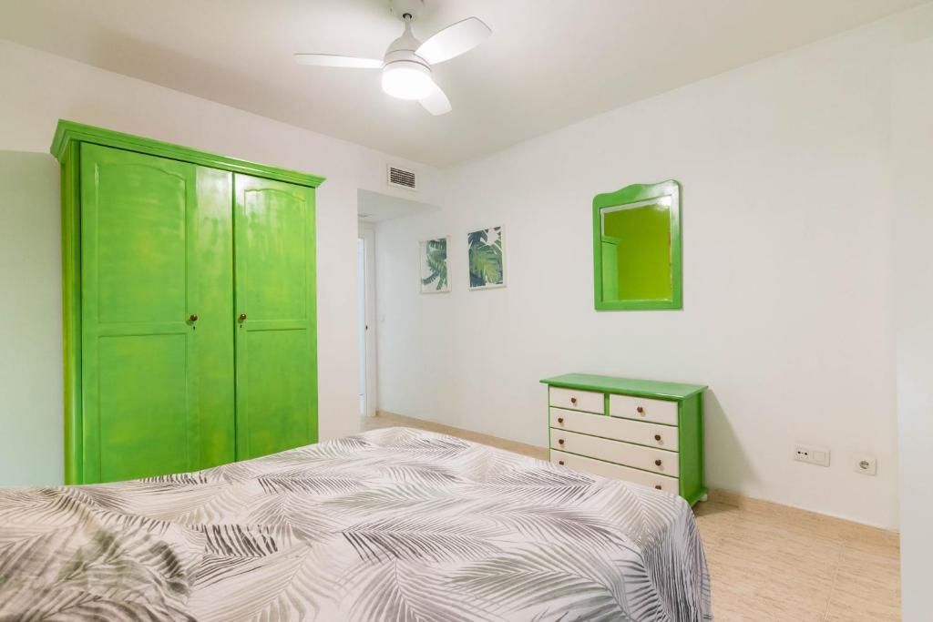 Dormitorio con armario verde, espejo y cómoda. Cama con funda de hojas. Paredes y techo blancos.