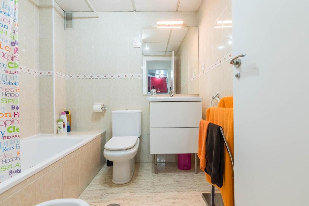 Baño con bañera, inodoro, lavabo y toallas. Paredes y suelo de color claro.