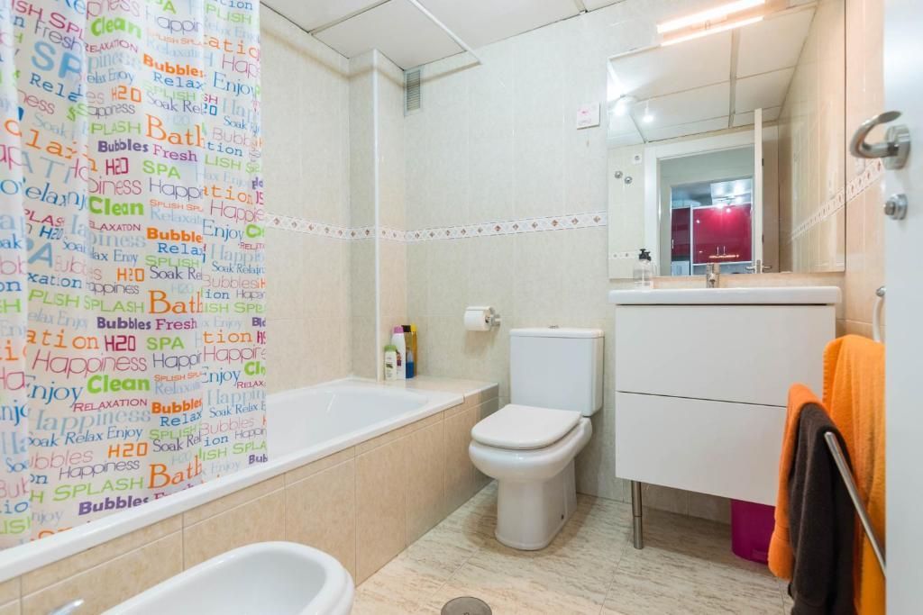 Baño con ducha, inodoro y lavabo. Paredes beige, grifería blanca y cortina de ducha colorida. Toallas naranjas y negras.