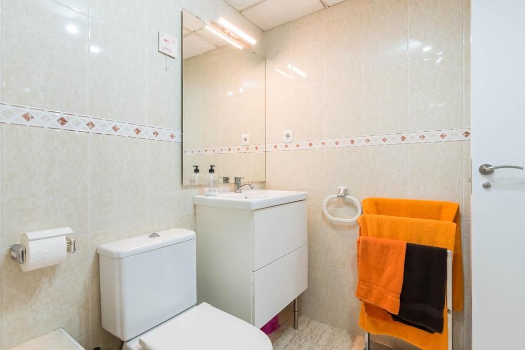 Pequeño baño con inodoro, lavabo y azulejos de color blanco; toallas de color naranja y negro en un estante.