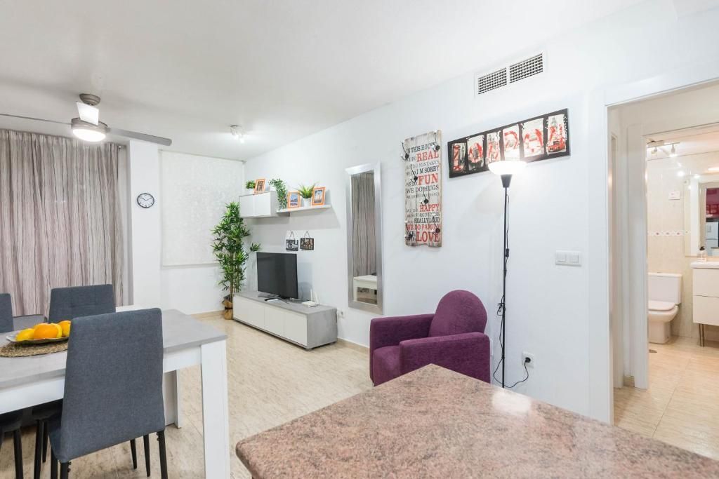Luminoso salón con mesa de comedor, TV, sillón morado y vista a un baño.