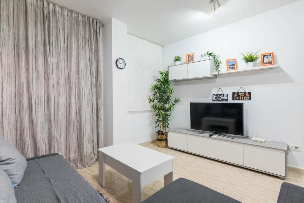 Sala de estar con sofá gris, mesa de centro blanca, TV y plantas decorativas.