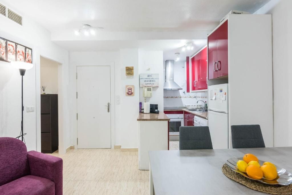 Sala de estar con un sofá morado, una mesa de comedor y una cocina con detalles en rojo visibles.
