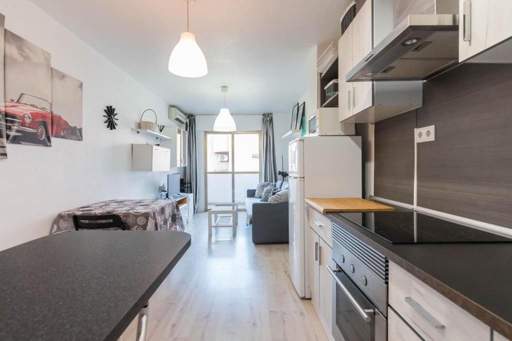 Interior de apartamento pequeño: cocina con encimeras, electrodomésticos blancos y sala de estar con sofá, balcón y cuadros.