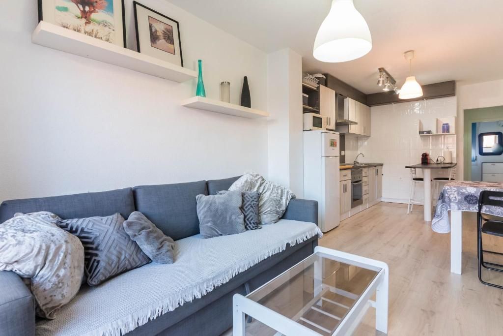 Sala de estar con sofá gris, cocina, estanterías con decoración y una mesa de centro de cristal.