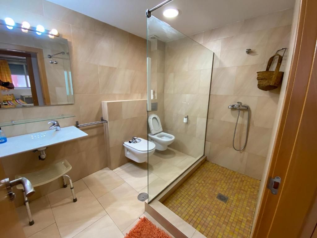 Baño con paredes de azulejos, ducha, inodoro y lavabo. La ducha tiene mampara de cristal y hay un taburete cerca del lavabo.